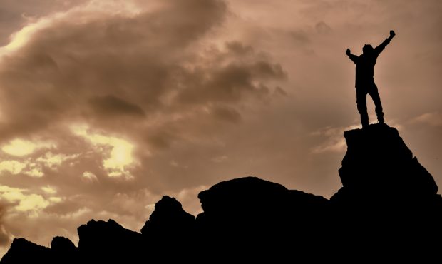 大きな岩の上でガッツポーズをする男性のシルエットと曇天