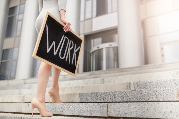 「WORK」と書かれた黒板を持って歩くハイヒールを履いた女性