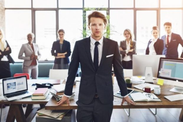 オフィスに立つ濃紺のスーツを着た男性と、背後に並ぶスーツ姿の人々
